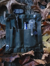 A waxed canvas EDC travel tray laid flat on a wooden surface with various slots and compartments, containing some example EDC items like a flashlight and multitool. The tray is primarily dark-colored.