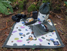 Outdoor camping gear laid out on a forest floor blanket, including a pair of black binoculars, a small black lantern, a dark green drawstring bag, a matching pouch, and a patterned picnic blanket with colorful mushroom and leaf motifs.