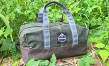 Green and brown duffel bag with a logo on a stone surface surrounded by greenery