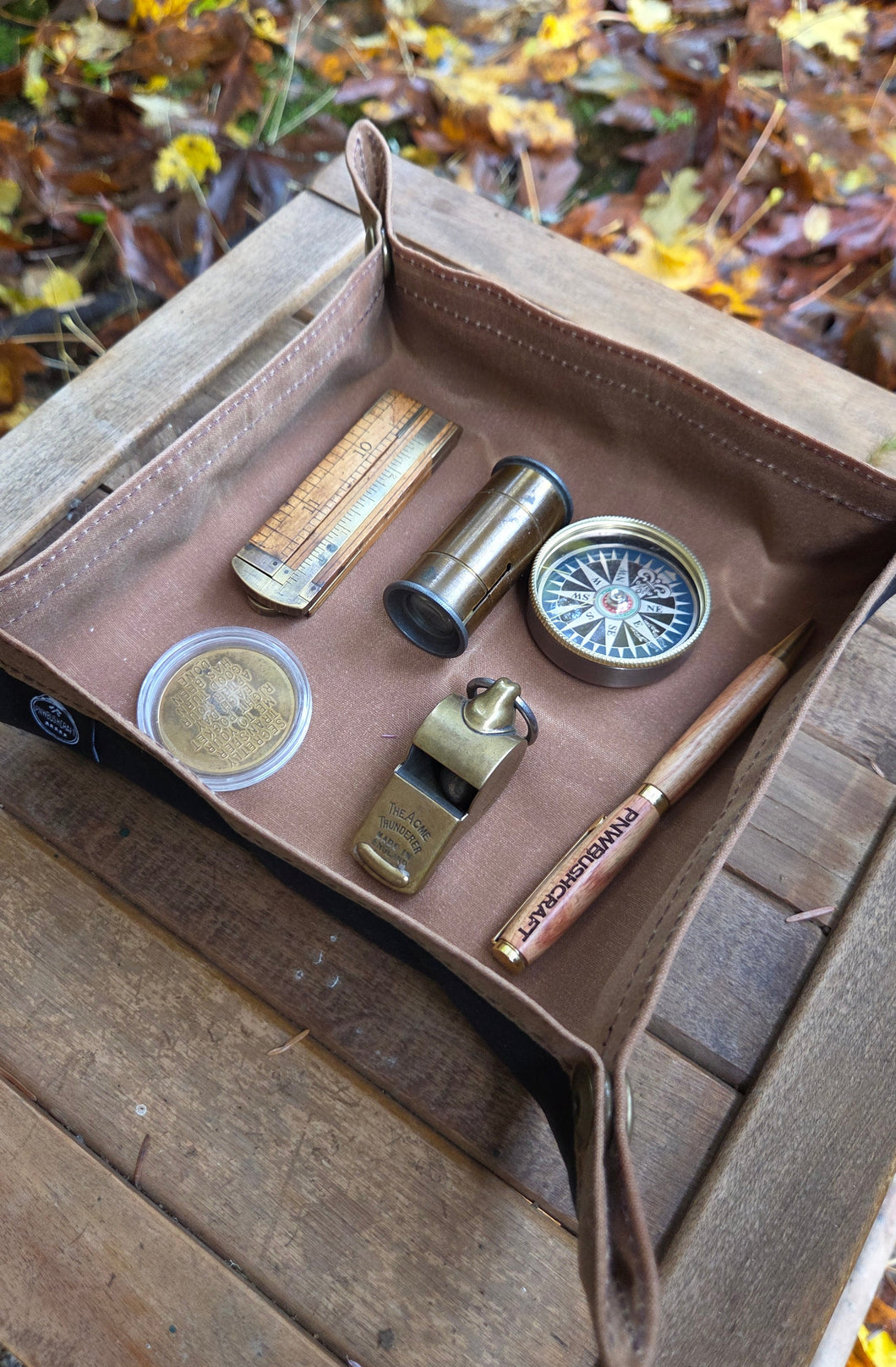 Collection of vintage items including a compass, pen, and coins in an open waxed canvas case on a wooden surface with a natural background.