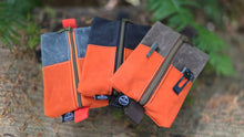 A stack of four zippered utility pouches in orange and gray canvas, photographed outdoors against a blurred natural background of green foliage and brown wood.