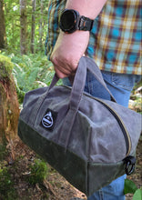A person outdoors in a forest holding a dark gray canvas utility bag with a top zipper and a small black-and-white logo patch on the front pocket.
