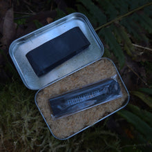 Two black rectangular objects in a small metal container on a green leafy background