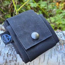 A black canvas utility pouch with a snap button closure, resting on a birch log outdoors with green foliage in the background.