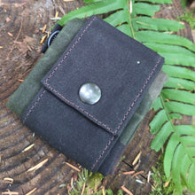 A black canvas utility pouch with a snap button closure, resting on a mossy log outdoors with green ferns in the background.