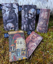 Four gothic-themed fabric card sleeves with printed illustrations of skulls, a witch, and tarot cards, displayed on moss and a birch log outdoors.