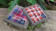 Two canvas utility pouches with plaid fabric inserts, displayed on a weathered wooden surface with green ferns in the background. One pouch features a blue, red, and purple plaid pattern, and the other features an orange, red, and white plaid pattern.