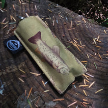A tan canvas pouch featuring a printed brown trout illustration, resting on a cut tree stump outdoors with pine needles and forest foliage visible.