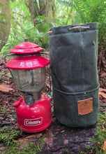 A red Coleman lantern standing next to a dark green canvas bag with a brown leather patch, set outdoors on a log in a forest with ferns and trees in the background.