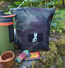 A camouflage pouch with a black hook-and-loop patch and white logo, shown outdoors on mossy rocks with green foliage in the background. A green bottle, a terracotta cup, and a small colorful item are partially visible at the bottom.